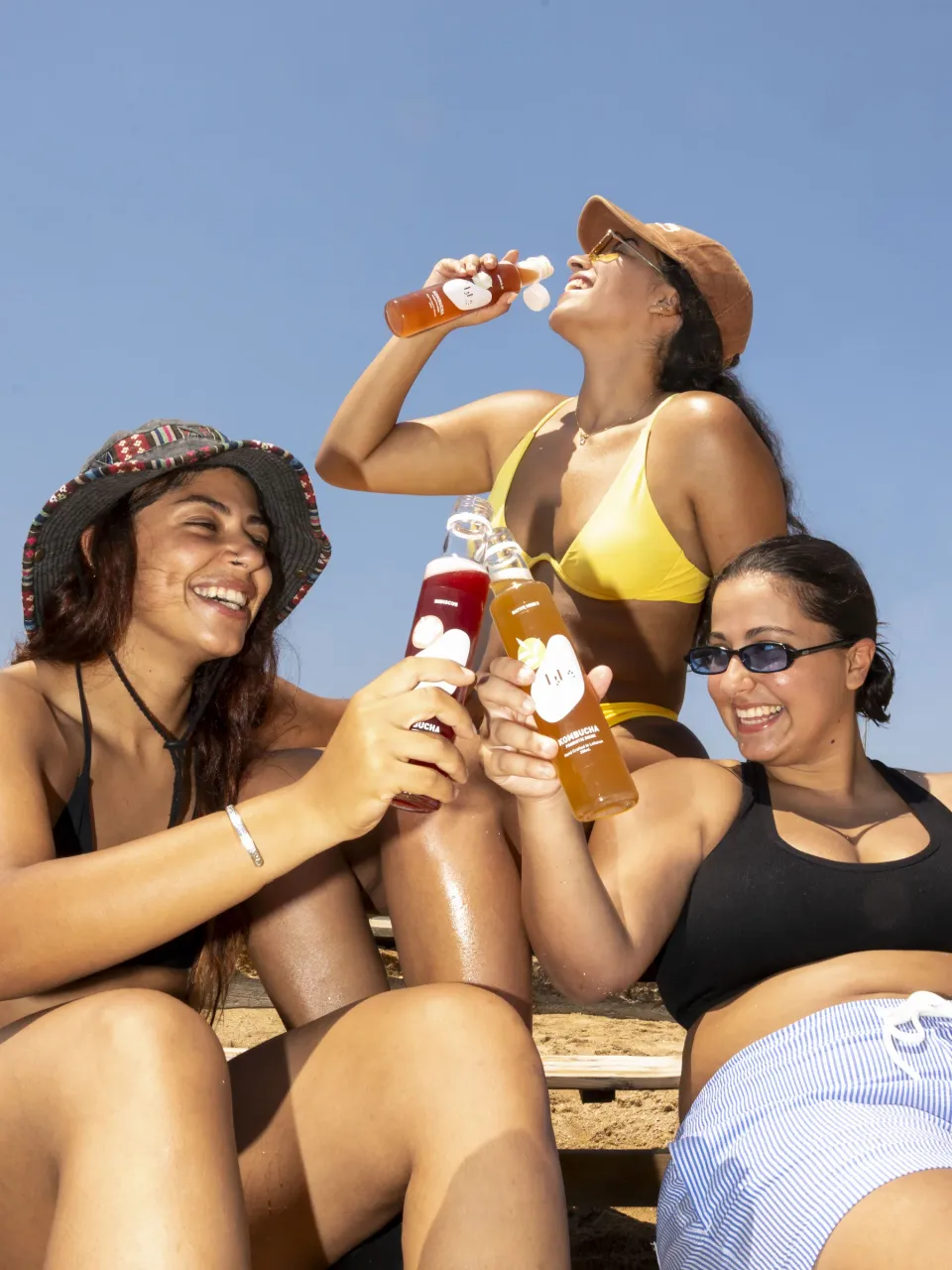 Three friends on a beach clinking Shaya kombucha bottles